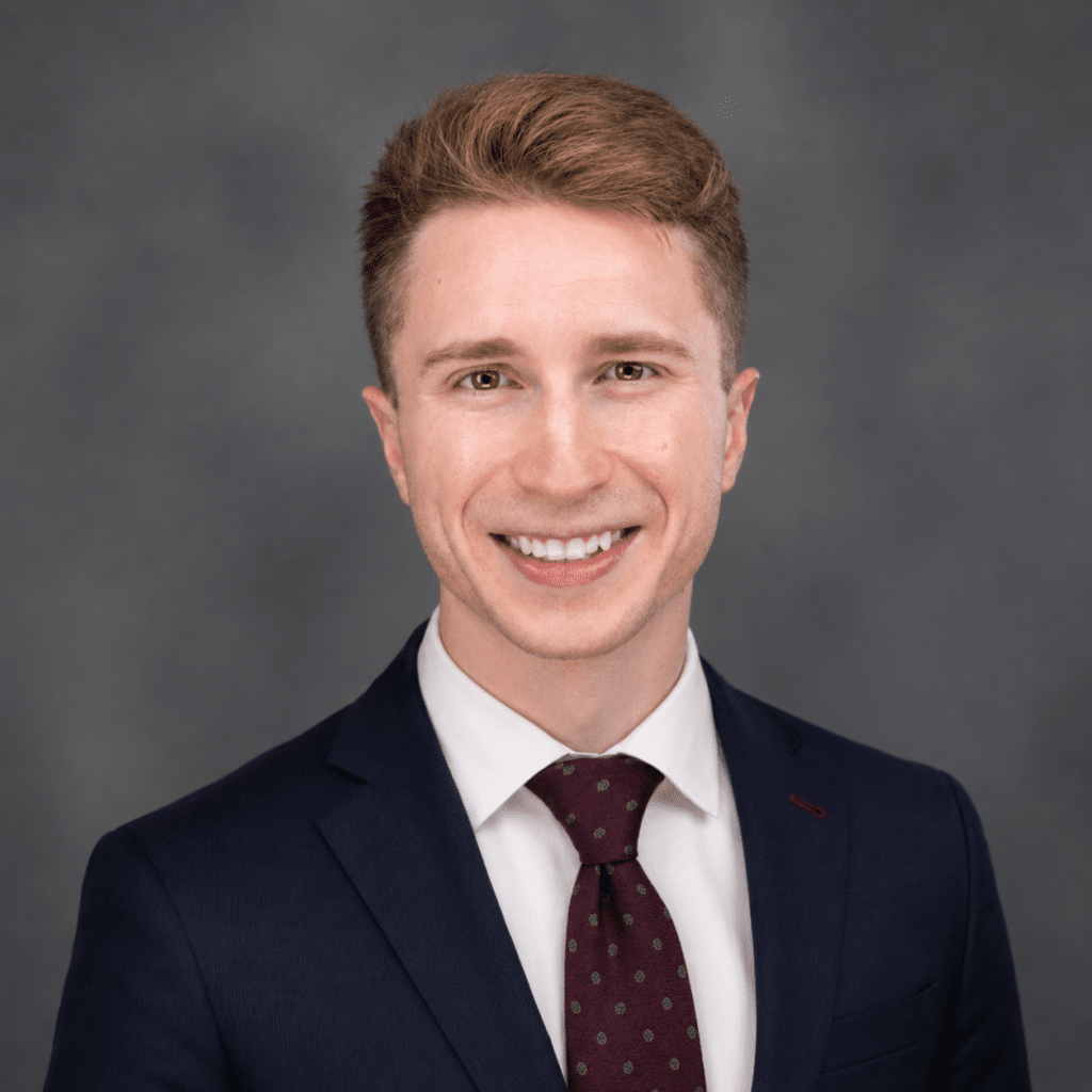 A young man with short light brown hair, wearing a dark navy suit, white shirt, and maroon polka dot tie, smiles in front of a gray studio backdrop.